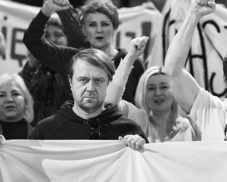 Czarno-białe zdjęcie. Tomasz Augustynowicz stoi na pierwszym planie wśród protestujących w teatrze. Trzyma przed sobą duży baner, a jego twarz ma skupiony wyraz. Za nim inne osoby unoszą zaciśnięte pięści w geście sprzeciwu.